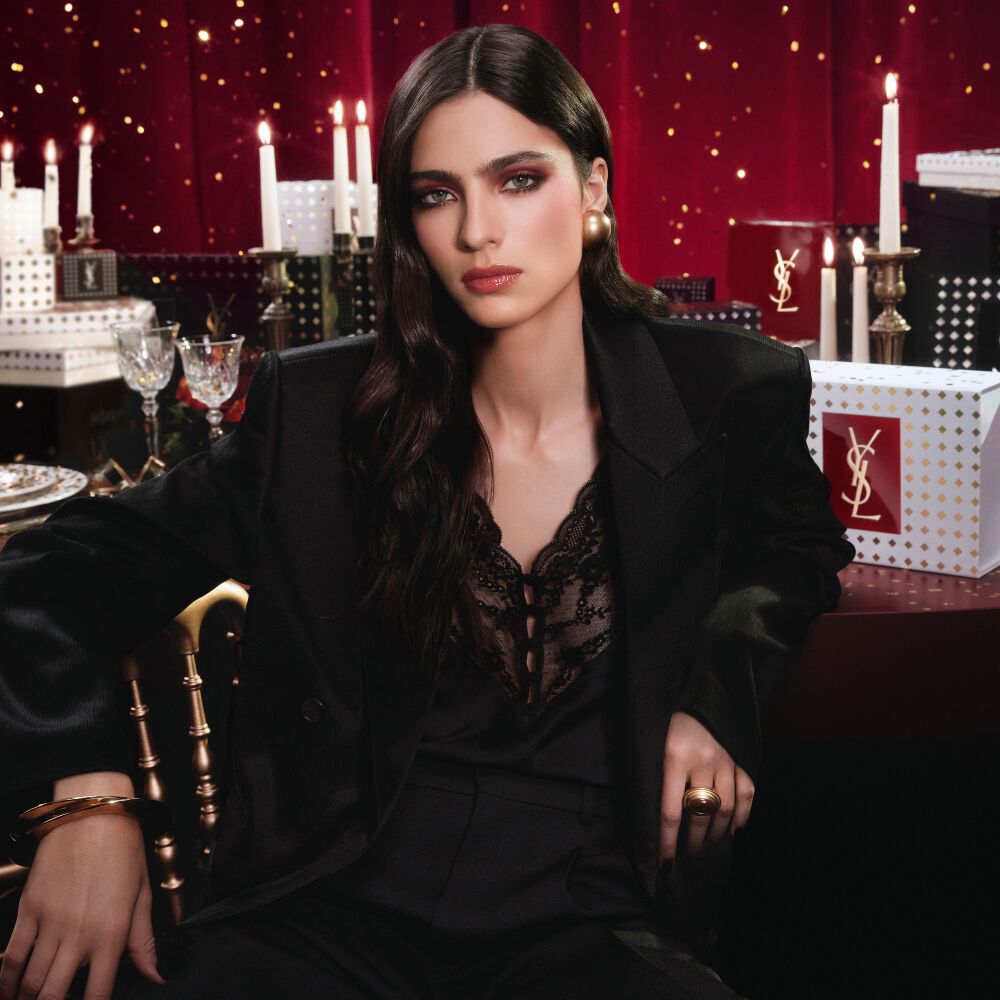Modelo vistiendo un traje negro y un look glamuroso, sentada en un ambiente festivo iluminado por velas con cajas de regalo de YSL. Modelo vistiendo un traje negro y un look glamuroso, sentada en un ambiente festivo iluminado por velas con cajas de regalo de YSL. Modelo vistiendo un traje negro y un look glamuroso, sentada en un ambiente festivo iluminado por velas con cajas de regalo de YSL. Banner B7 oculto.