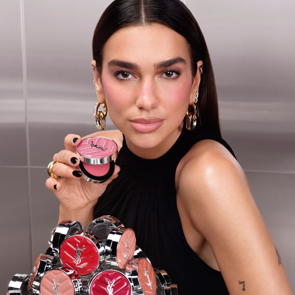 Dua Lipa sostiene un estuche abierto de YSL Make Me Blush Lifted Glow Cream Blush, con una coleccion de los nuevos tonos desplegados frente a ella.