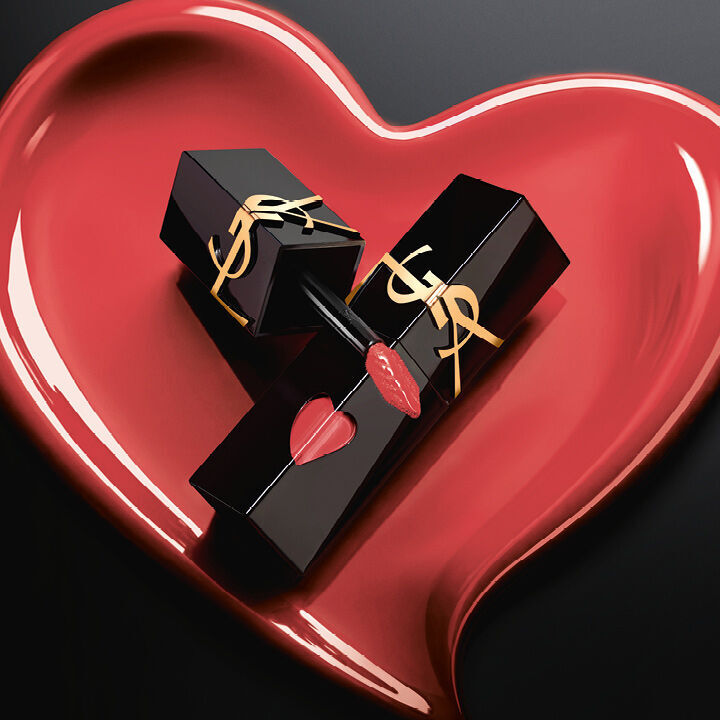 Dos frascos de tinta labial YSL en negro mate con recortes de corazon rosas y logos dorados, exhibidos dentro de un corazon esculpido en rosa mate a juego sobre fondo negro.