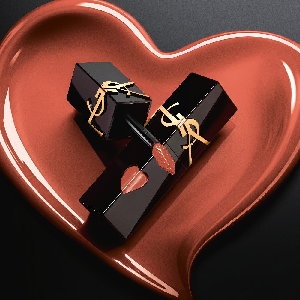 Dos frascos de tinta labial YSL en negro mate con recortes de corazon rosas y logos dorados, exhibidos dentro de un corazon esculpido en rosa mate a juego sobre fondo negro.