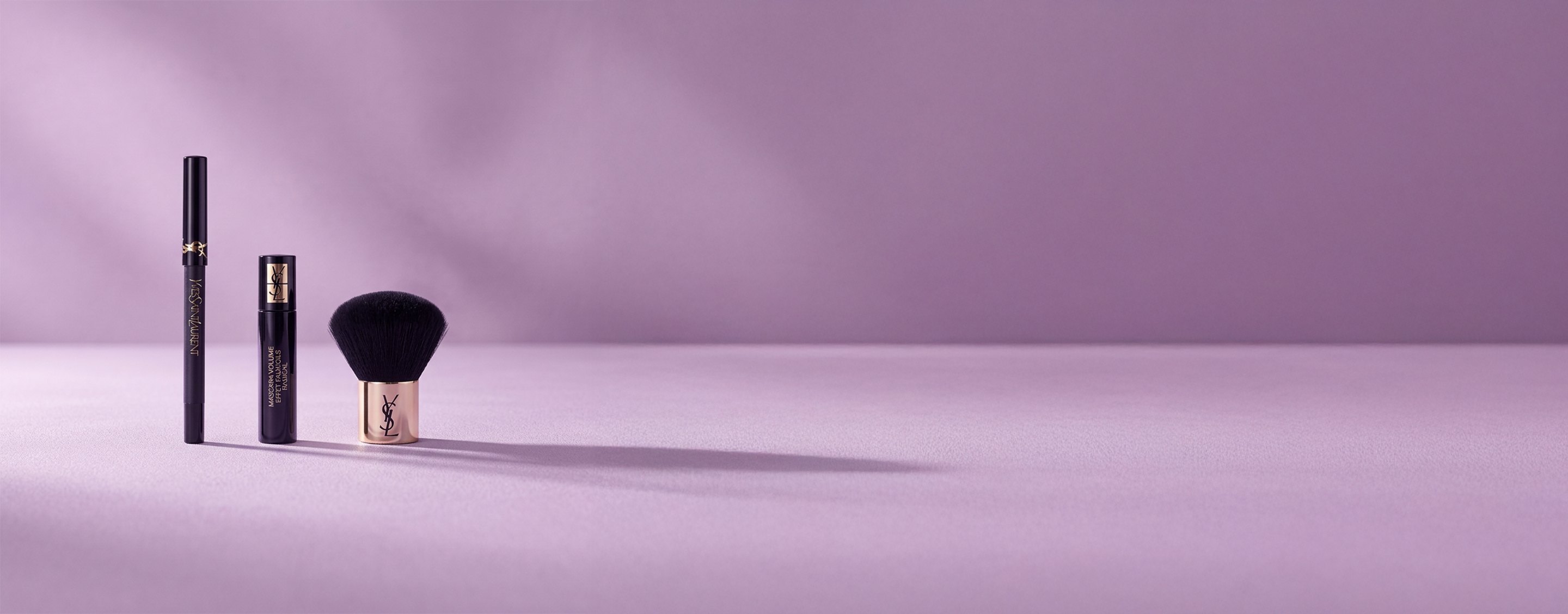 Tres productos de belleza de Yves Saint Laurent, incluyendo una máscara de pestañas y una brocha kabuki, sobre un fondo lavanda suave.