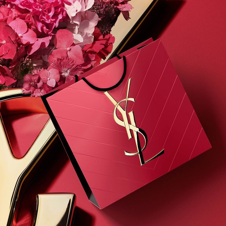 Caja de regalo rosa de YSL con el icónico logotipo Cassandre dorado, rodeado de pétalos de flores primaverales.