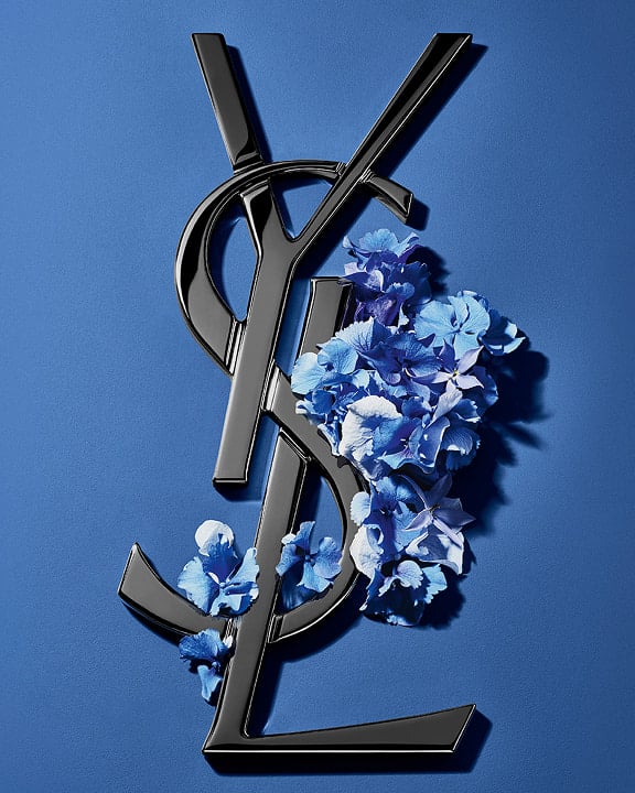 Logotipo Cassandre de YSL en negro, adornado con vibrantes flores primaverales azules, sobre un fondo azul sereno.