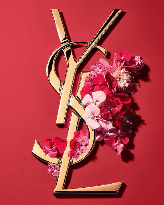 Logotipo Cassandre dorado de YSL, adornado con vibrantes flores rosas primaverales, sobre un fondo rosa.