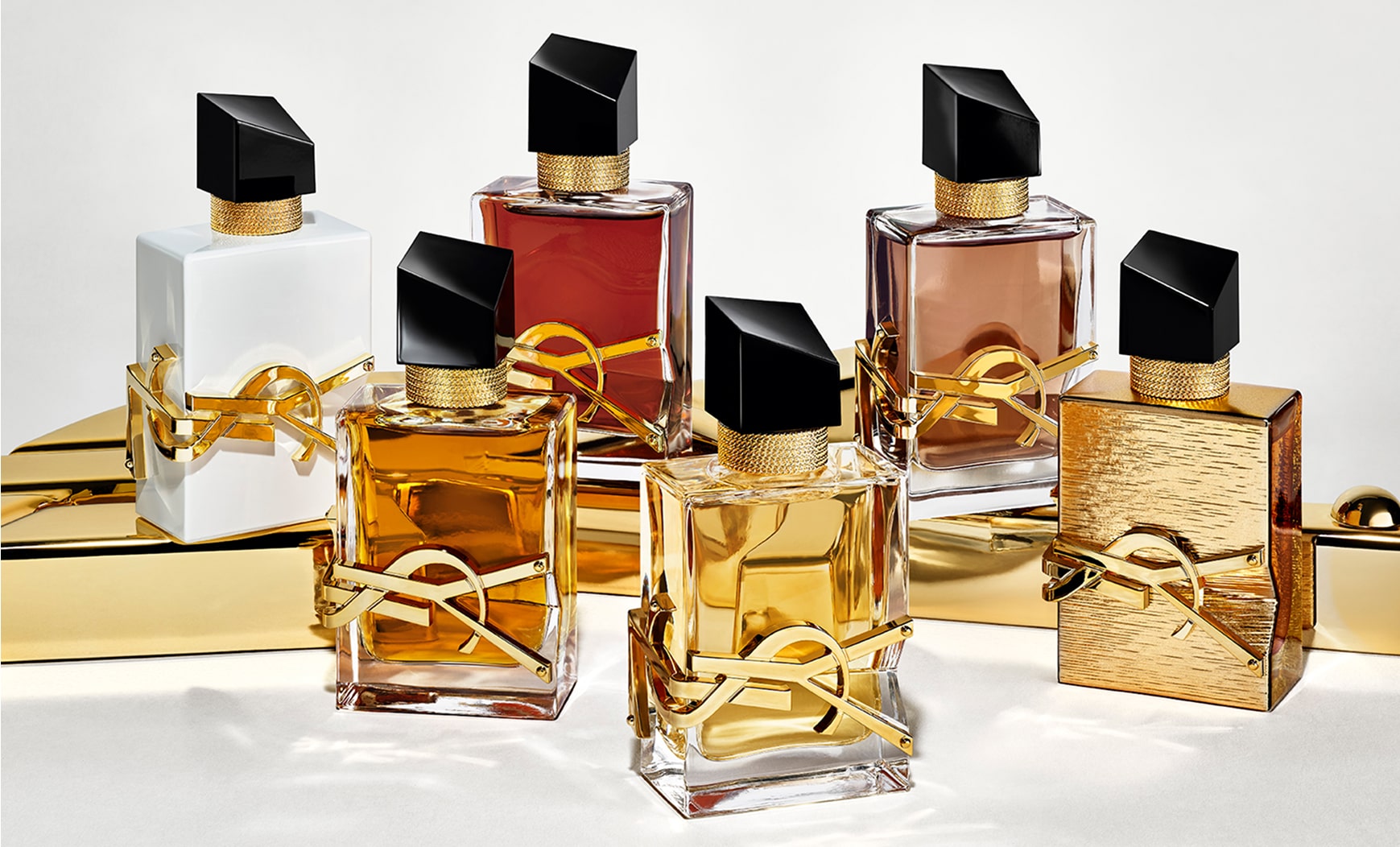 Dos fragancias del perfume YSL Libre se presentan sobre una superficie dorada brillante. Los frascos presentan variaciones en el color del líquido y el acabado, incluyendo tonos transparentes, blancos, rosados ​​y marrones, y un frasco con textura completamente dorada. Todos lucen las icónicas letras YSL en dorado y una fuente negra inclinada.

