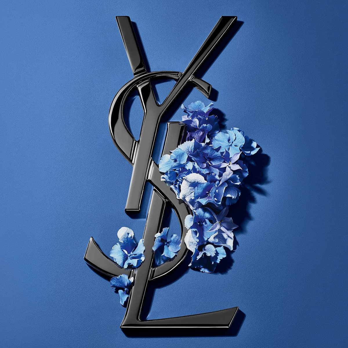Logotipo Cassandre de YSL en negro, adornado con vibrantes flores primaverales azules, sobre un fondo azul sereno.