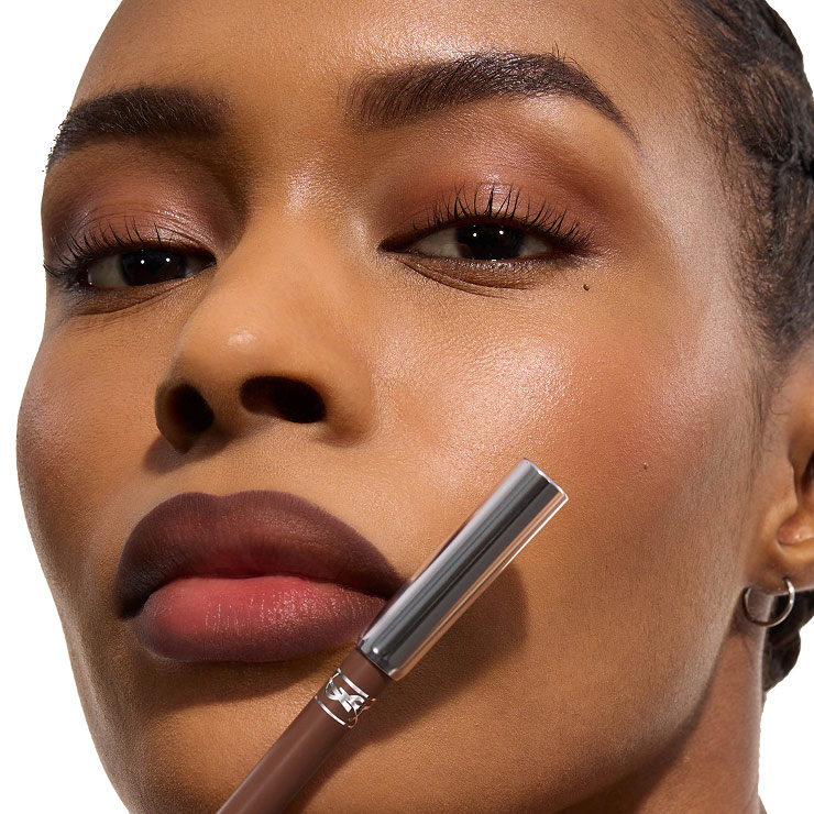 Primer plano de una modelo con labios degradados en color marrón sosteniendo un perfilador YSL a juego.
