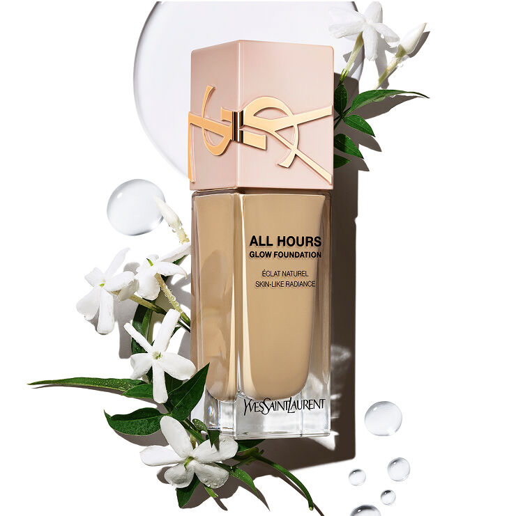 Frasco de la base de maquillaje YSL All Hours Glow con 'Éclat Naturel Skin-Like Radiance' rodeado de flores de jazmín blancas y gotas de agua claras, sugiriendo ingredientes naturales e hidratación.