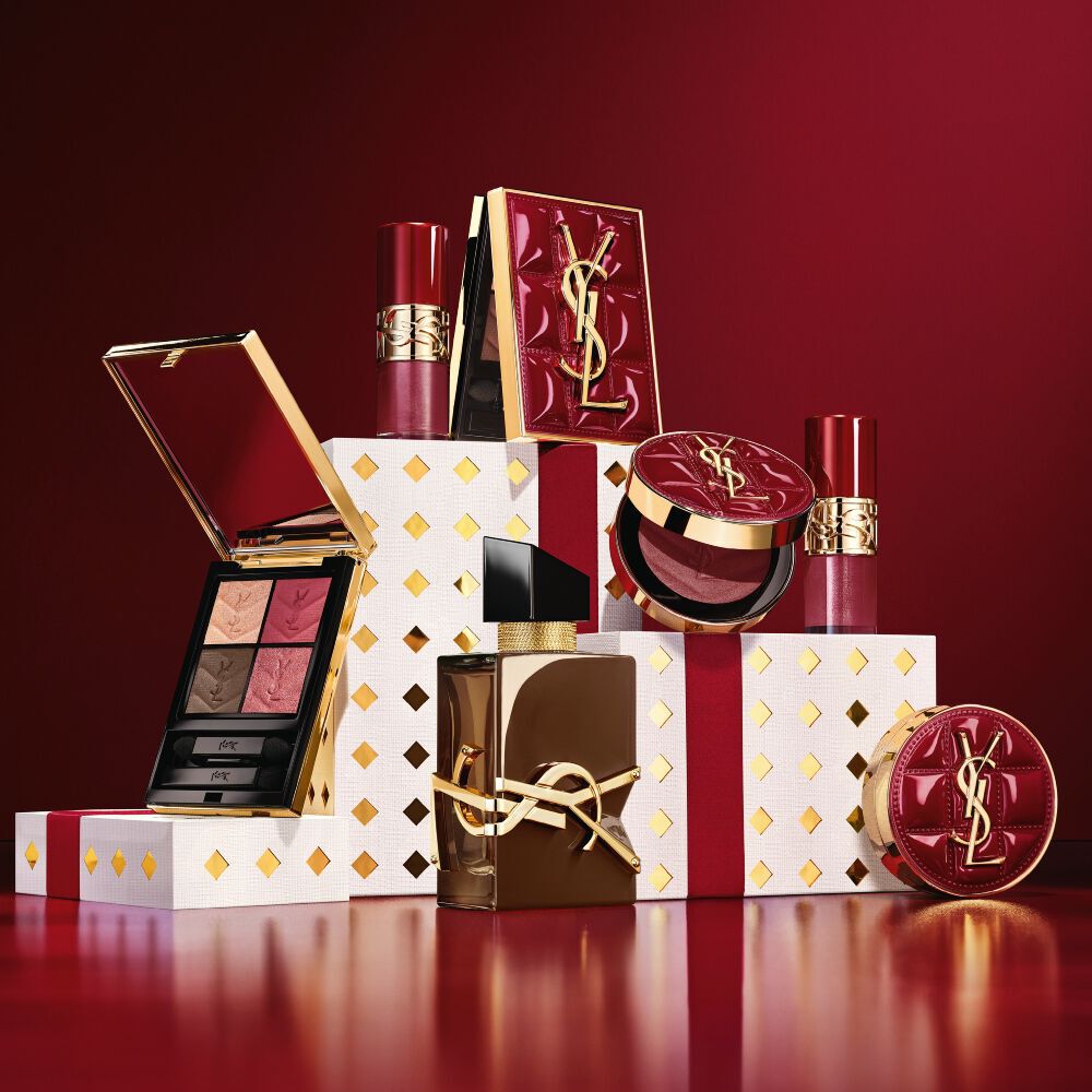 Colección de maquillaje y cofre regalo de fragancias YSL Navidad 2026 sobre pedestales blancos con puntos dorados en burdeos.