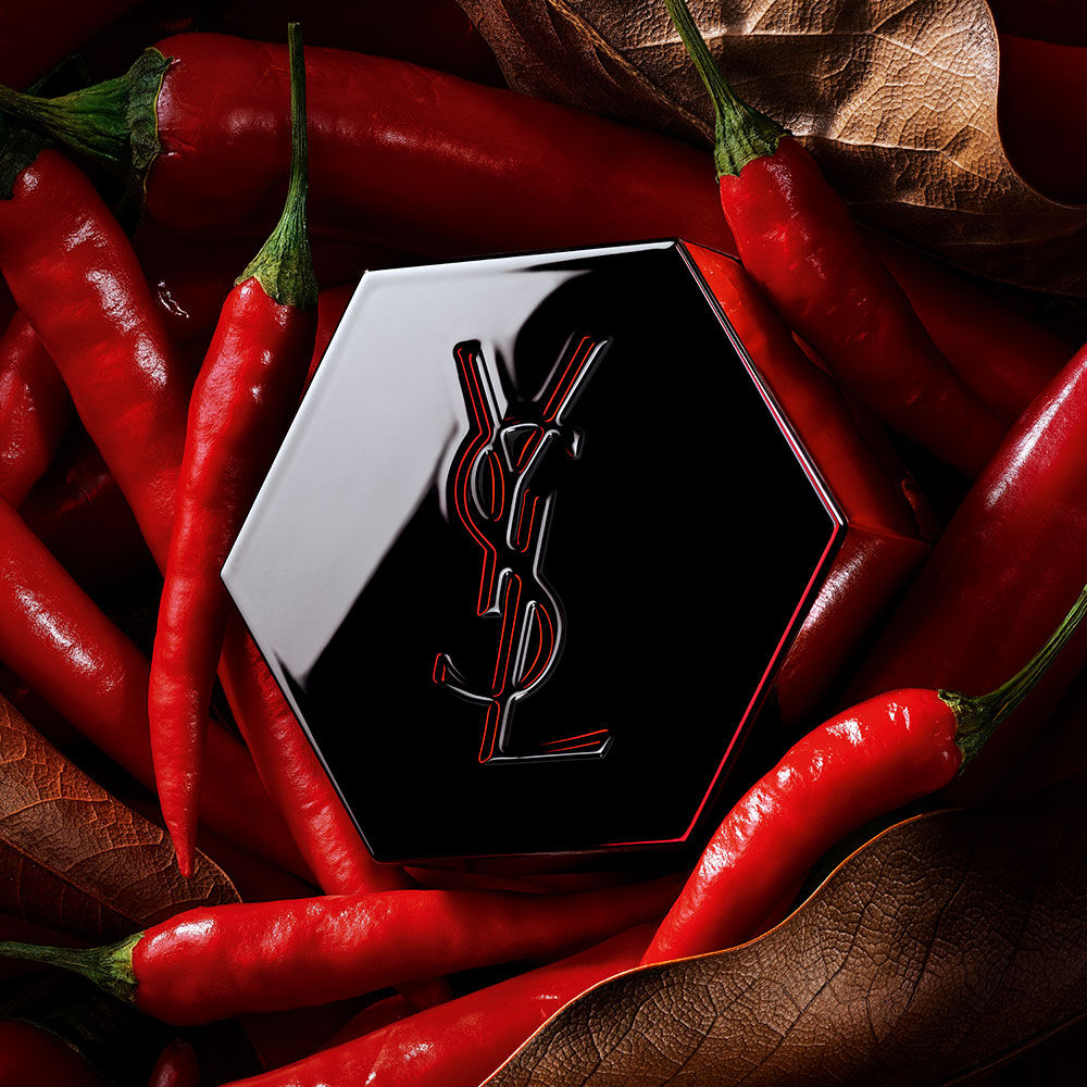Tapón de fragancia hexagonal negro de YSL con el logotipo perfilado en rojo, rodeado de chiles rojos.
