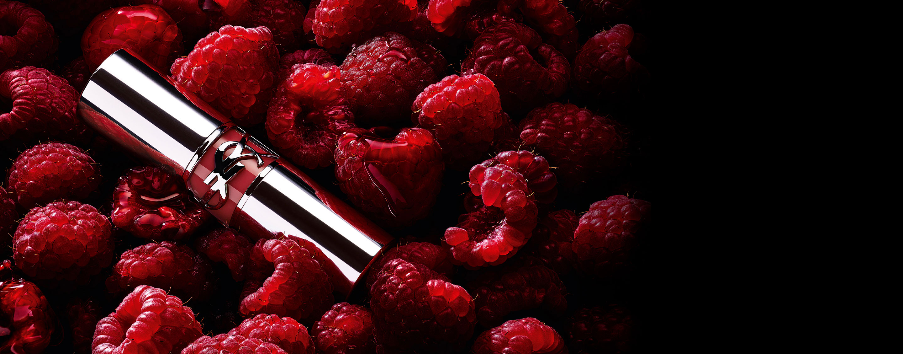 Barra de labios YSL LOVESHINE Berry Crush, en un envase plateado con el logotipo de YSL en color baya, situada entre frambuesas frescas.