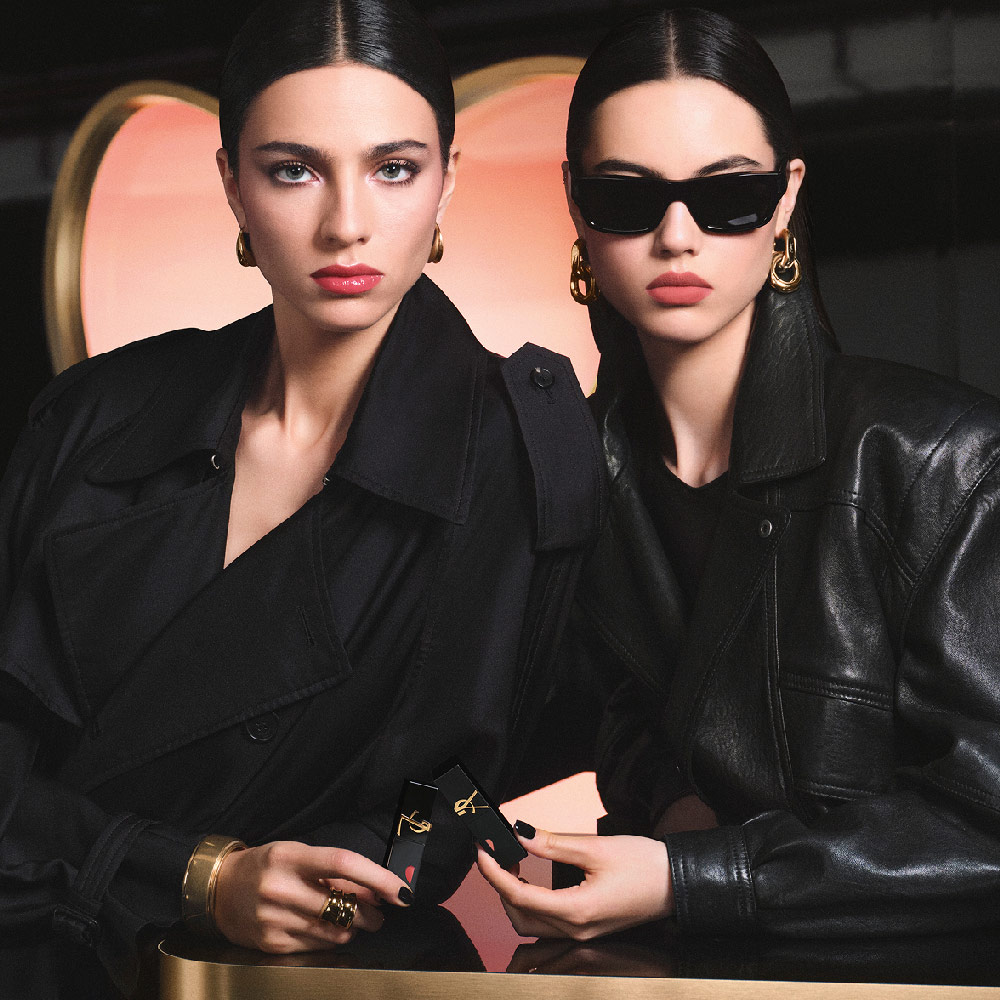 Dos modelos chic con gabardina negra y chaqueta de cuero sosteniendo labiales YSL frente a un gran fondo rosa en forma de corazón, luciendo un look de labios rojos brillantes y otro de rosa mate suave.