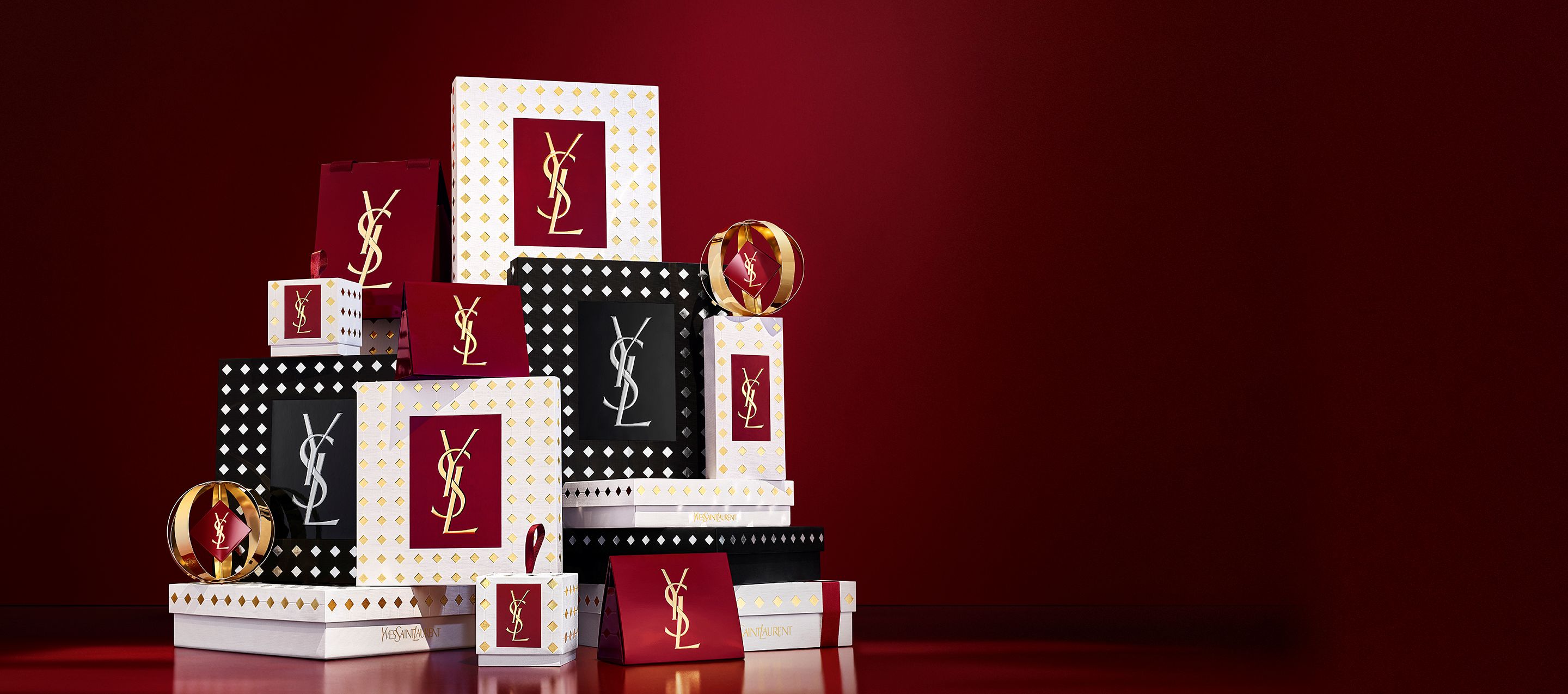 Tres modelos con elegantes outfits negros sostienen cajas regalo de YSL frente a un telón rojo festivo con confeti dorado cayendo.
