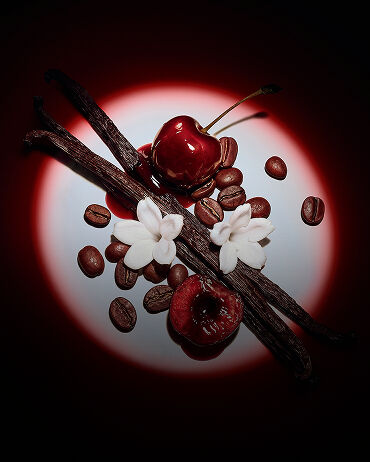 Primer plano de jugosas cerezas rojas, flores de jazmín, vainas de vainilla y granos de café, realzados por un dramático foco para Black Opium Eau de Parfum Over Red.
