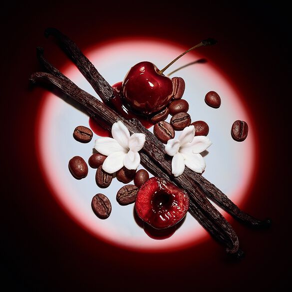 Primer plano de jugosas cerezas rojas, flores de jazmín, vainas de vainilla y granos de café realzados por un foco dramático para Black Opium Eau de Parfum Over Red.