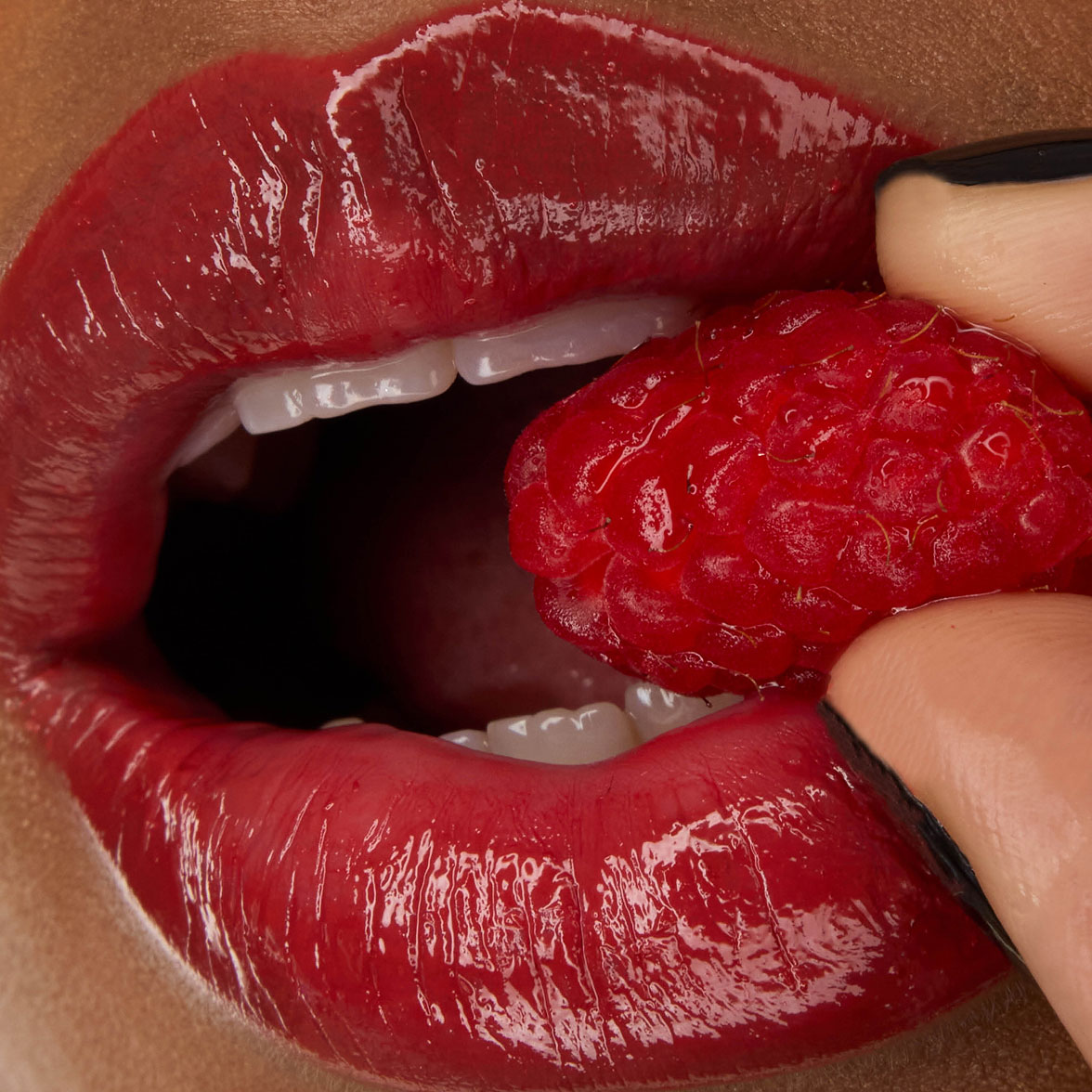 Primer plano de los labios de la modelo Zahara, realzados con la barra de labios YSL LOVESHINE en el tono Berry Crush 219, sosteniendo una frambuesa fresca.