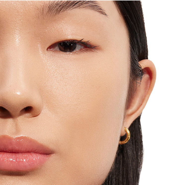 Primer plano detallado del rostro de una persona, centrado en la zona de los ojos y las mejillas con maquillaje de aspecto natural y un pendiente de aro dorado, demostrando la luminosidad de la piel.