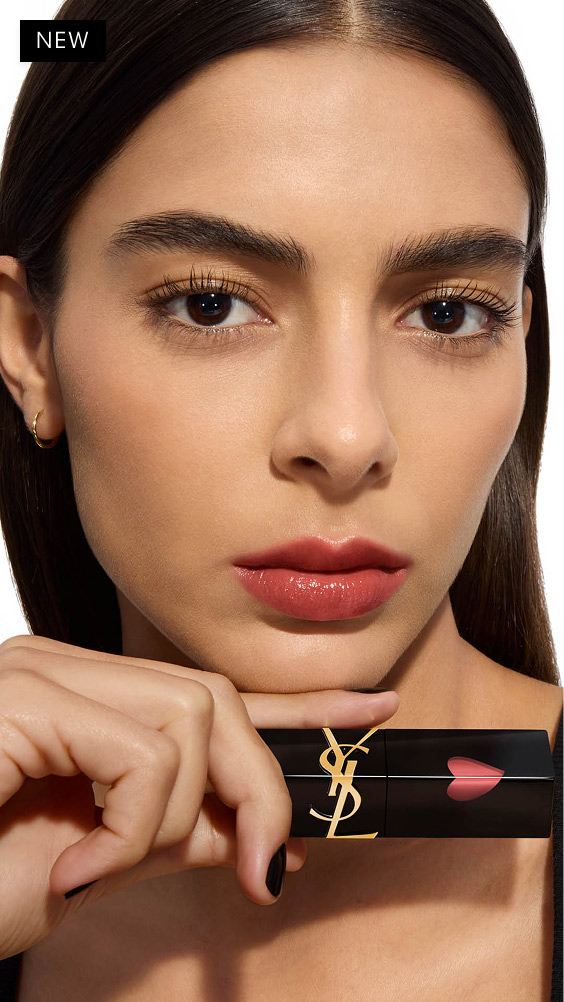 Modelo de tono de piel medio posando con la mancha labial YSL THE INKS VINYL CREAM sostenida horizontalmente bajo su mentón, mostrando un tono rosa cálido y brillante.