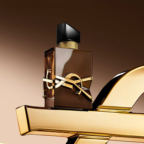 Frasco Libre Santal Couture marrón glaseado que refleja la luz, posado sobre una gran Cassandre YSL dorada.