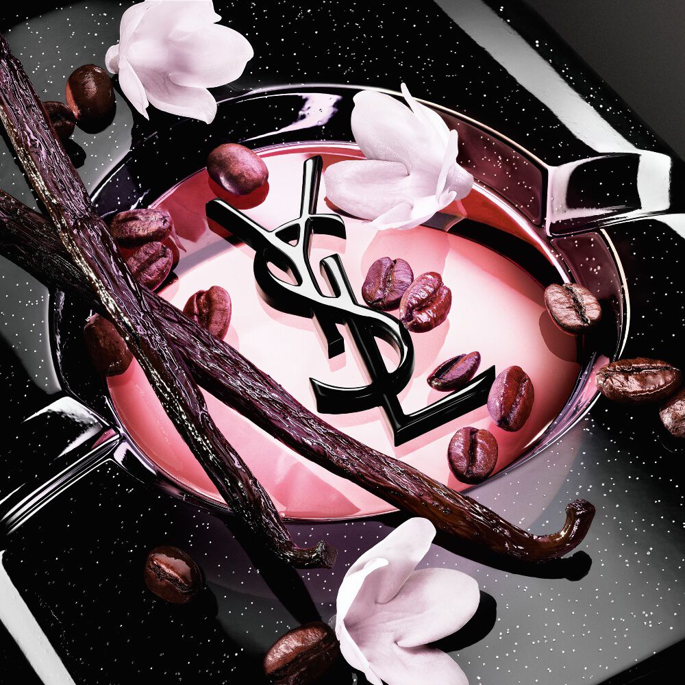 Close-up of roasted coffee beans, vanilla pods, and jasmine flowers arranged over the iconic Black Opium Eau de Parfum  bottle with the YSL Cassandre.