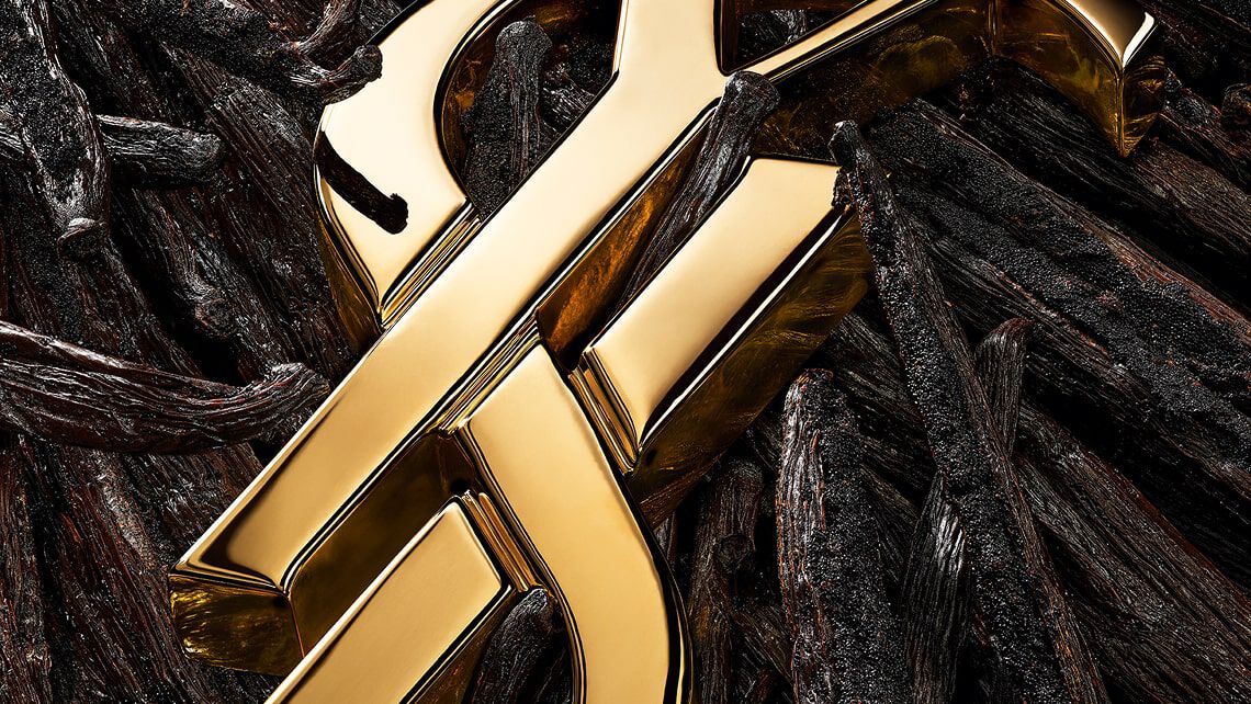 Primer plano del logo YSL dorado colocado entre vainas de vainilla, flores blancas y toques dorados brillantes.