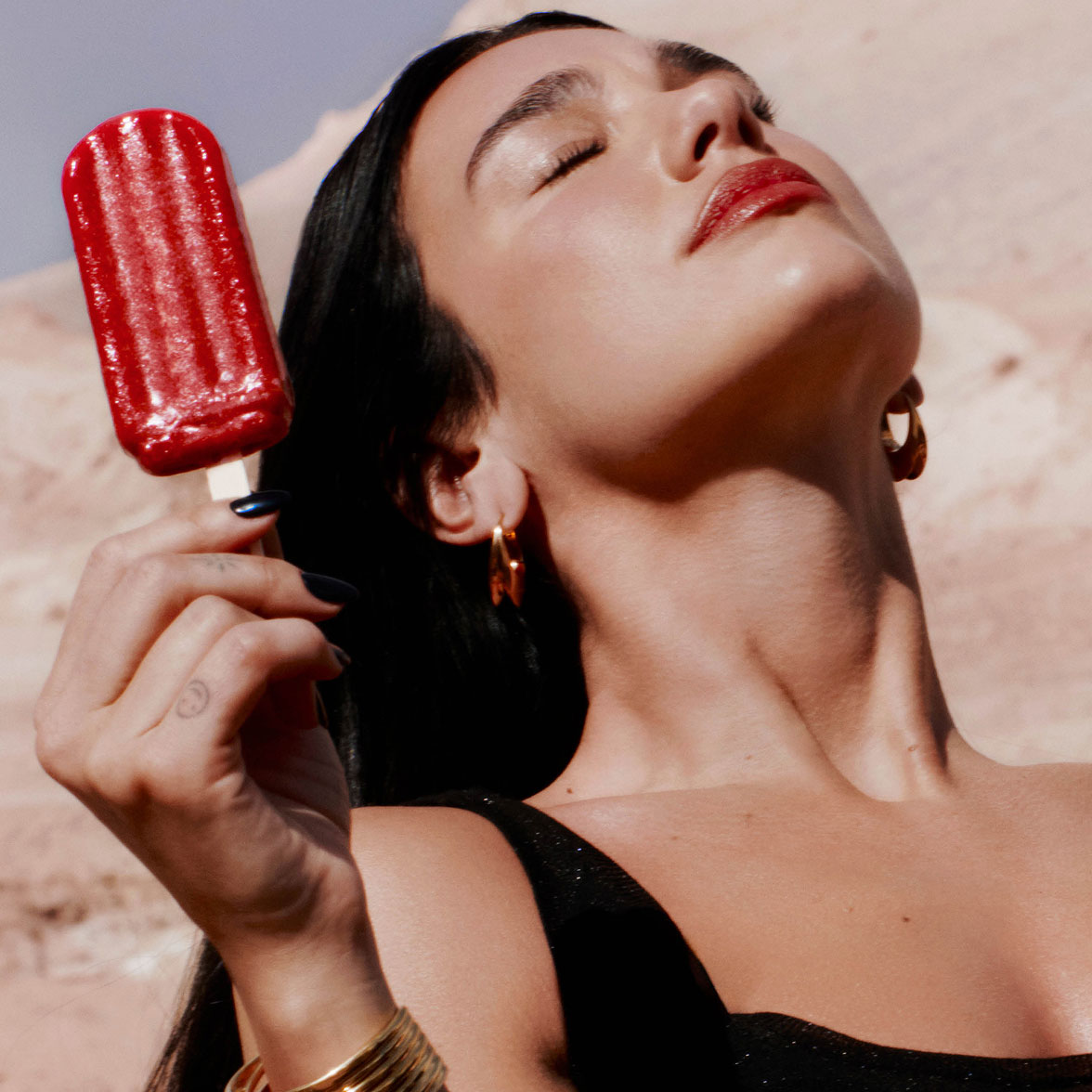 Un primer plano de Dua Lipa con los ojos cerrados bajo el sol, sosteniendo un polo de hielo rojo.