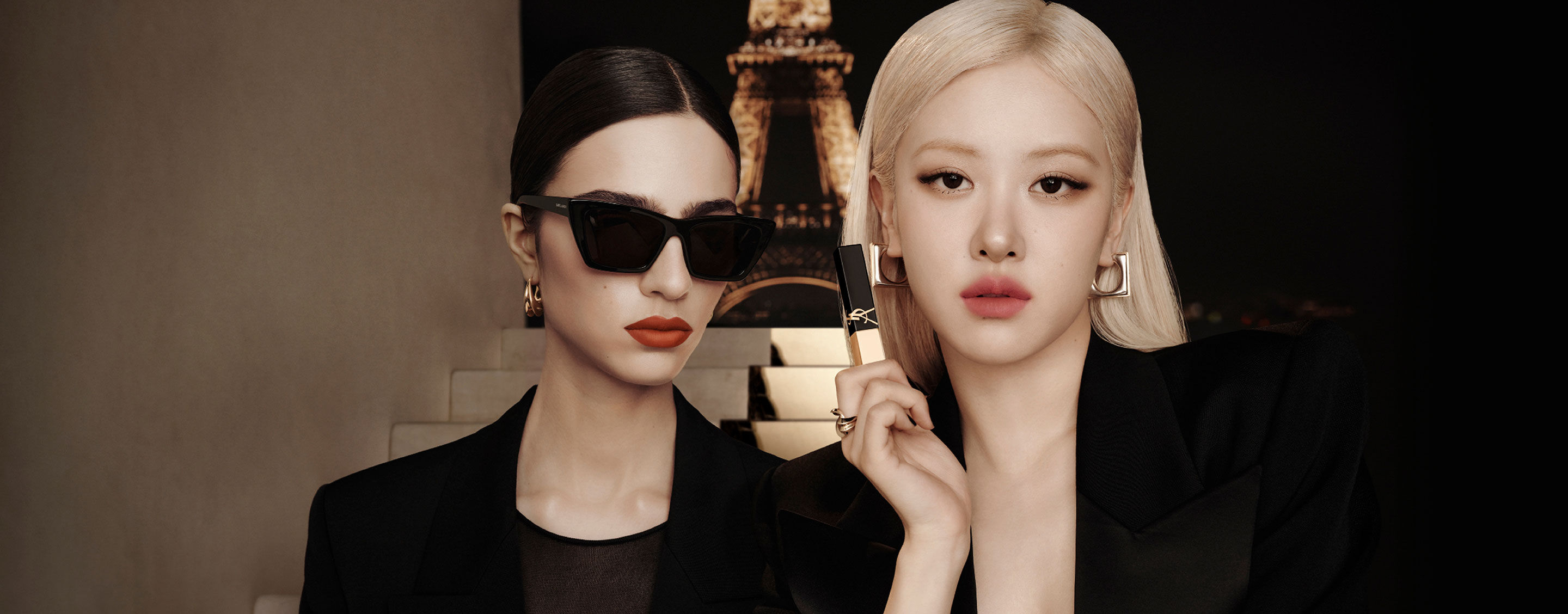 Dos modelos de negro frente a la Torre Eiffel de noche; una sostiene un labial YSL negro‑dorado.