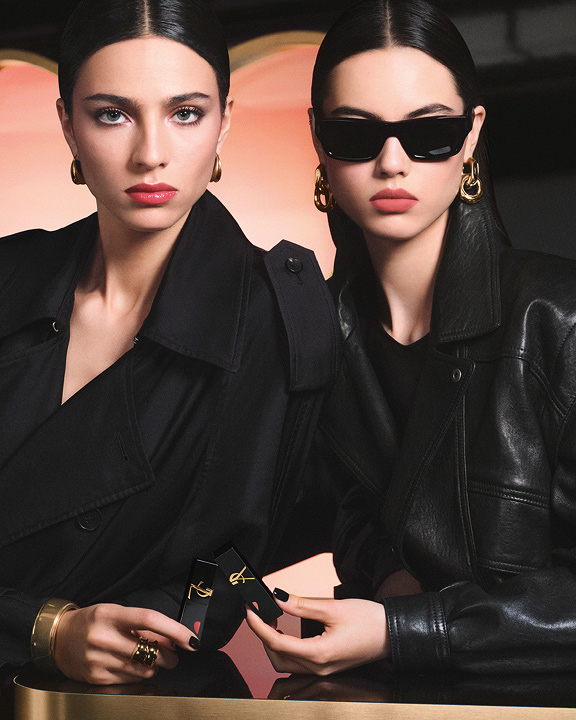 Dos modelos chic con gabardina negra y chaqueta de cuero sosteniendo labiales YSL frente a un gran fondo rosa en forma de corazón, luciendo un look de labios rojos brillantes y otro de rosa mate suave.