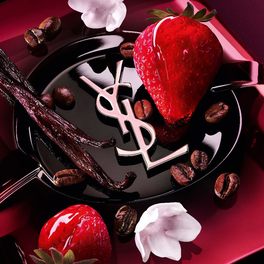 Primer
                plano de fresas frescas, vainas de vainilla, flores de jazmín y granos de café
                tostado dispuestos sobre el frasco de Black Opium Eau de Parfum Pink Glaze con el
                icónico cassandre de YSL.