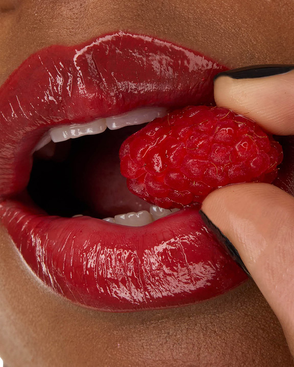 Primer plano de los labios de la modelo Zahara, realzados con la barra de labios YSL LOVESHINE en el tono Berry Crush 219, sosteniendo una frambuesa fresca.