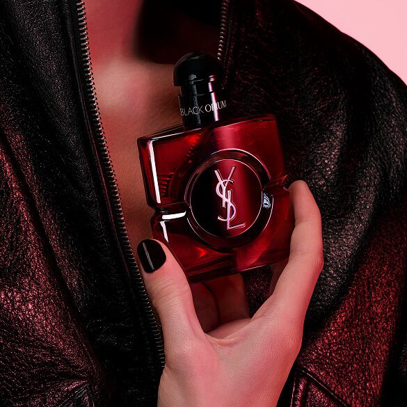 Primer plano de una mano con uñas negras sosteniendo el frasco Black Opium Eau de Parfum Over Red contra una chaqueta de cuero negra.