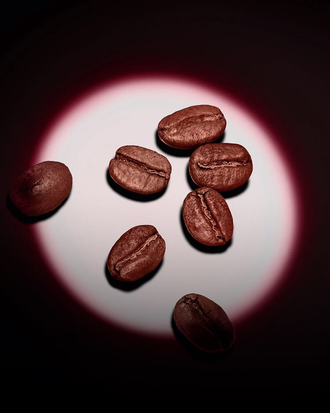 Un grupo de granos de café tostados destacados por un foco circular rosa sobre un fondo oscuro. 