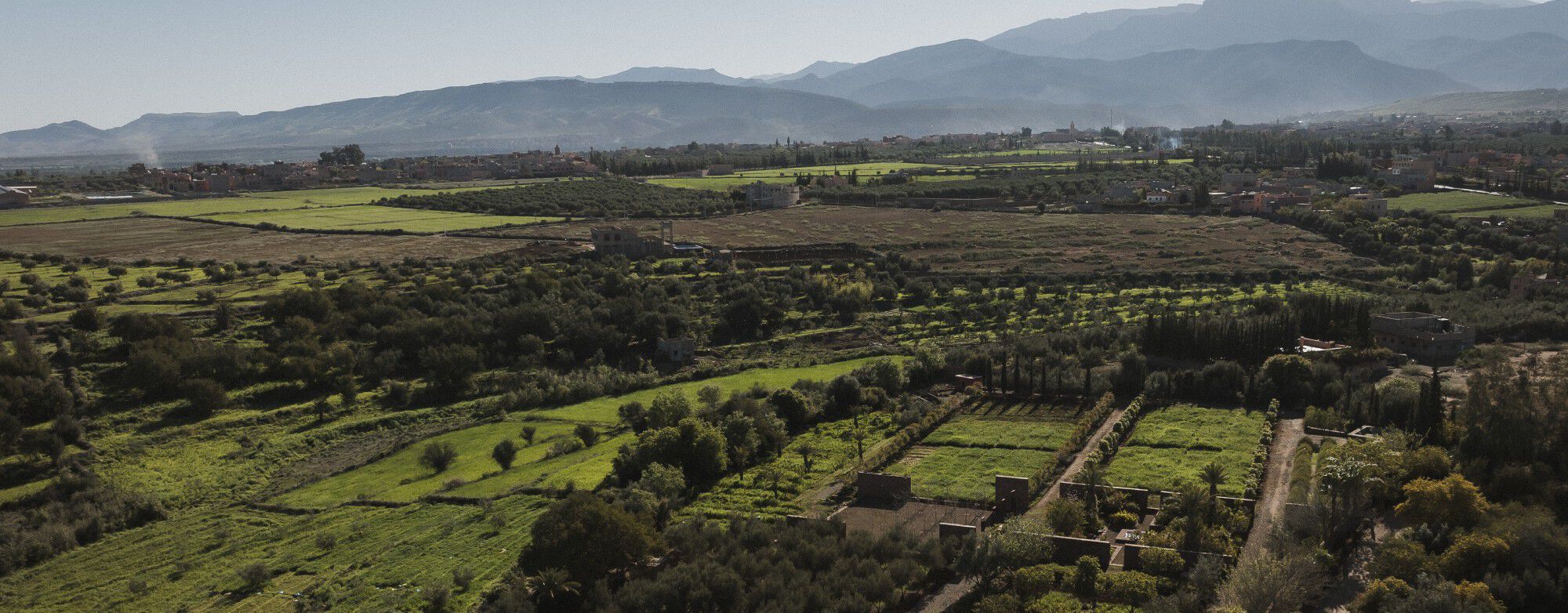YSL For The Future: Vista aérea de los Jardines Comunitarios del Ourika, con campos verdes exuberantes, árboles y montañas bajo un cielo despejado.