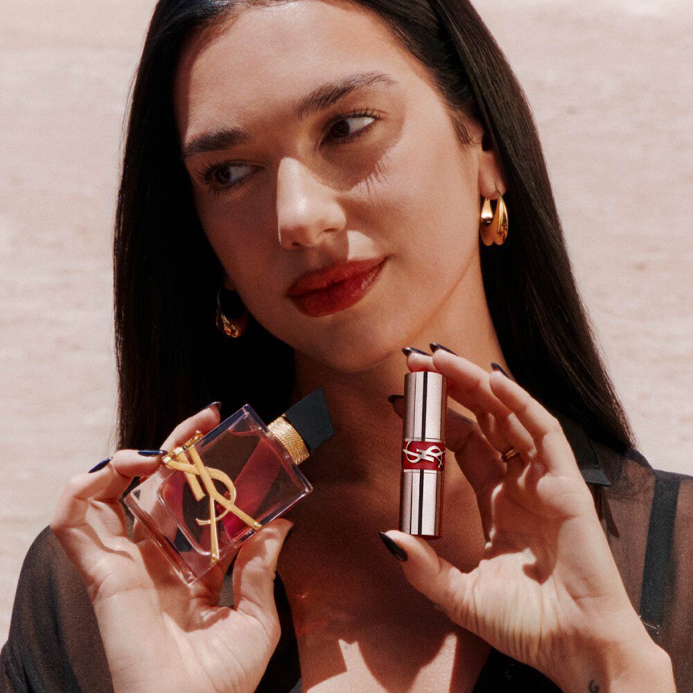 Dua Lipa sostiene Libre Eau de Parfum Berry Crush y un YSL Loveshine, con labios rojos y pendientes dorados.
